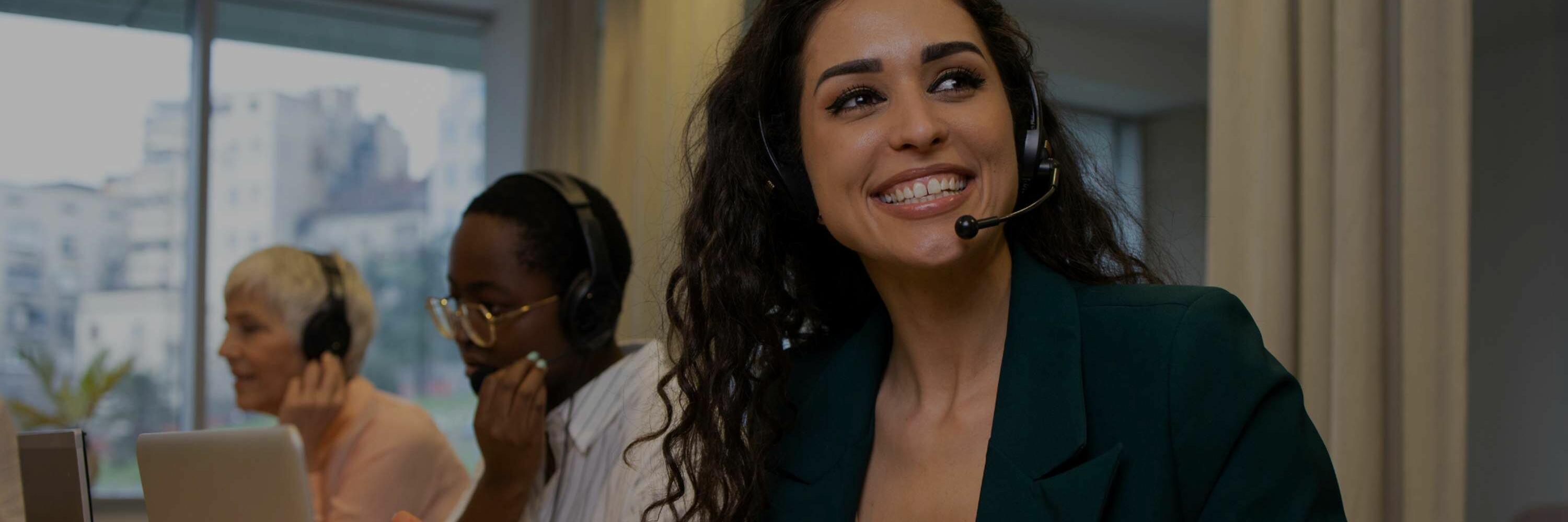 A woman with dark hair wearing a headset and smiling. Two call center employees are seated to her right.