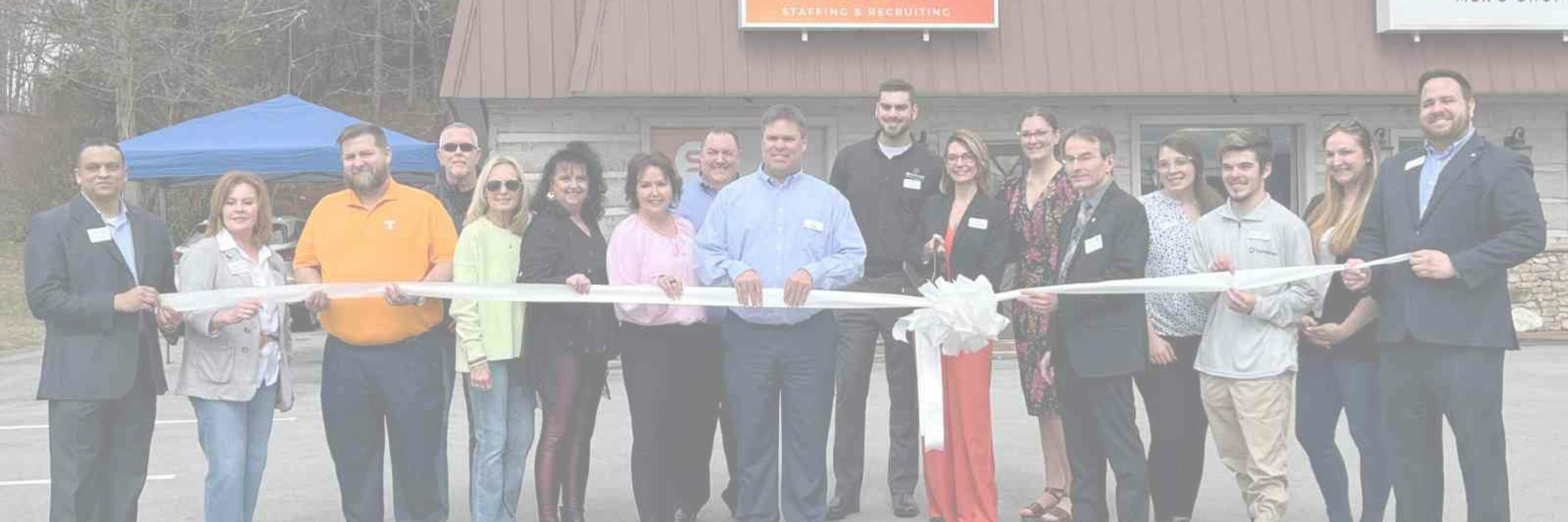 A group of people standing outside of a brown office building with an orange sign onthe roof that reads, Spherion Staffing & Recruiting. They are all holding up a white ribbon.