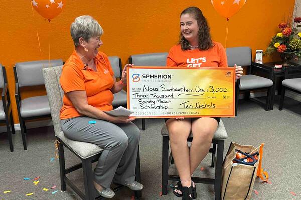 Two women in orange shirts sitting while holding a giant check