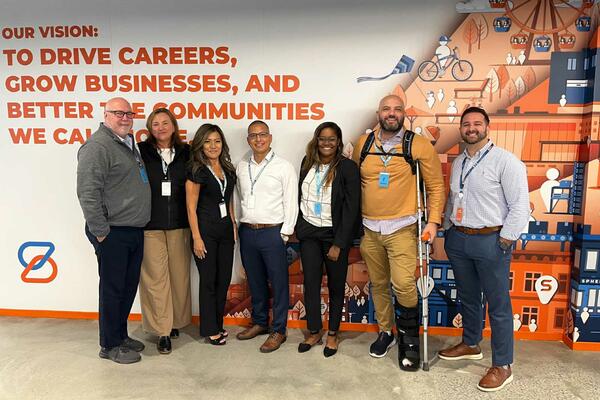 Photo of a group of smiling people standing in front of a white, orange and blue wall mnural