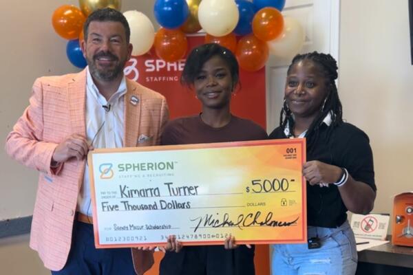 Three people smiling and standing behind a giant orange check in an office setting