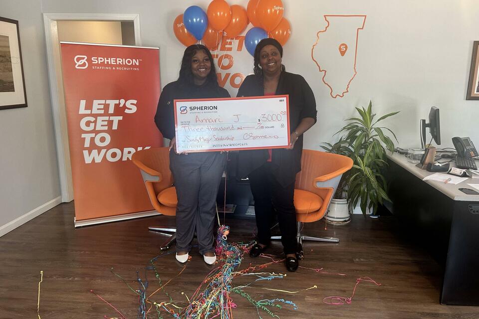 Two women holding a giant scholarship check in front of an orange banner that reads Let's Get to Work