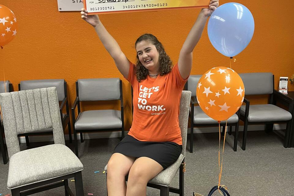 A young woman holding a giant check above her head in an orange room