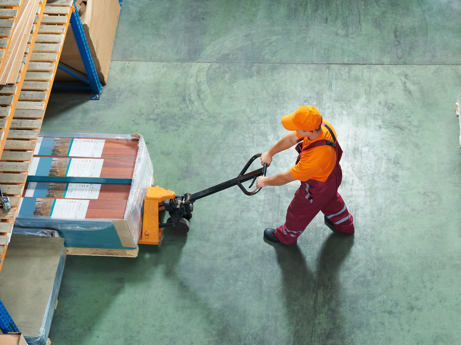 Birds'-eye view of a man in red overalls and a yellow hard hat pushed a pallet of goods into a space in a warehouse.