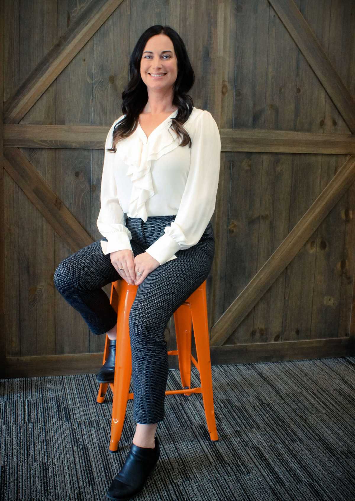 A woman in a white short and blue jeans sitting on an orange stool in front of a wooden wall