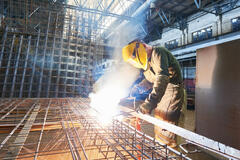 Welder wearing a yellow protective mask and using a heat tool to create a metal grid in a warehouse setting.