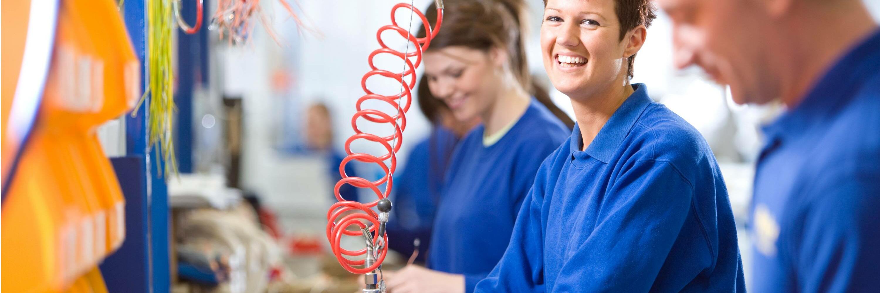 A woman in a blue work suit smiling while working with a red coil in a factory